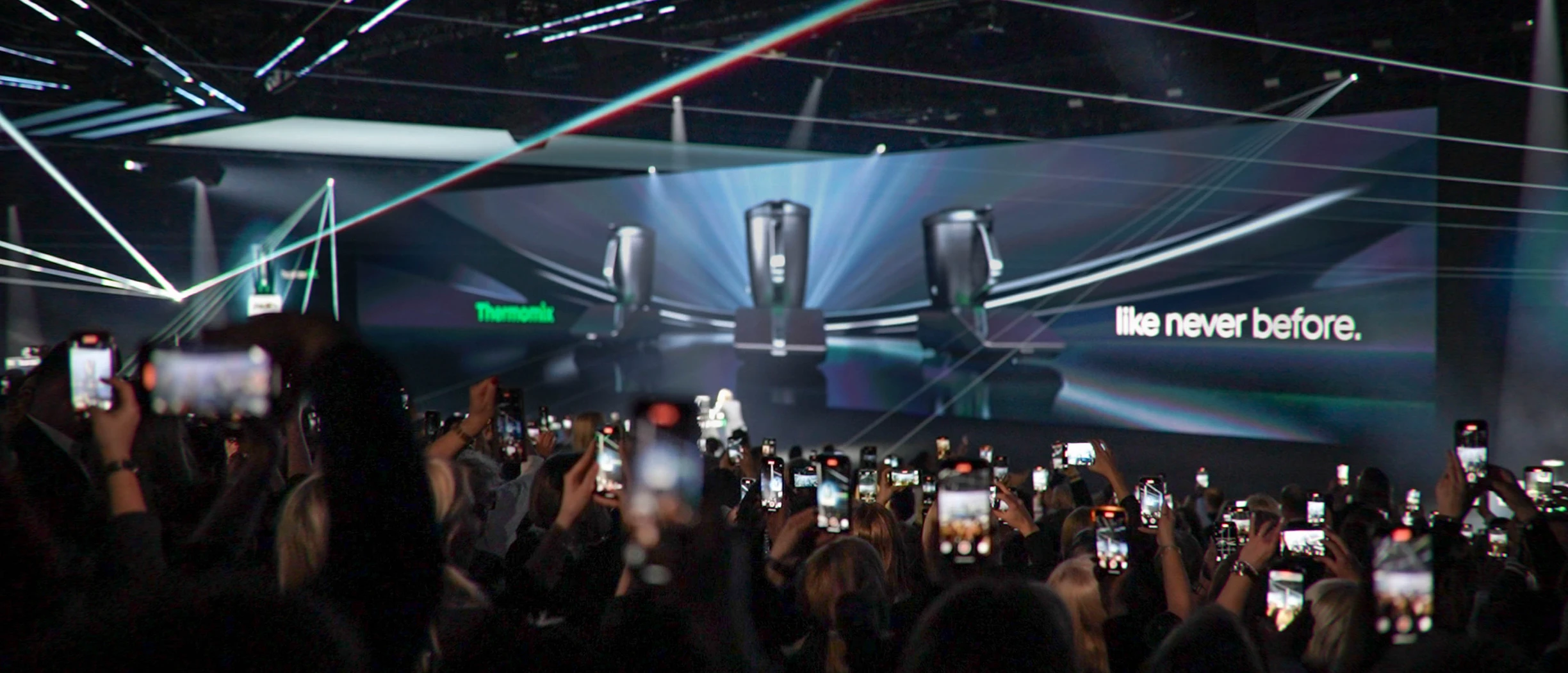Thermomix Produktpräsentation mit begeistertem Publikum und Smartphones. Neue Geräte auf großer Leinwand.